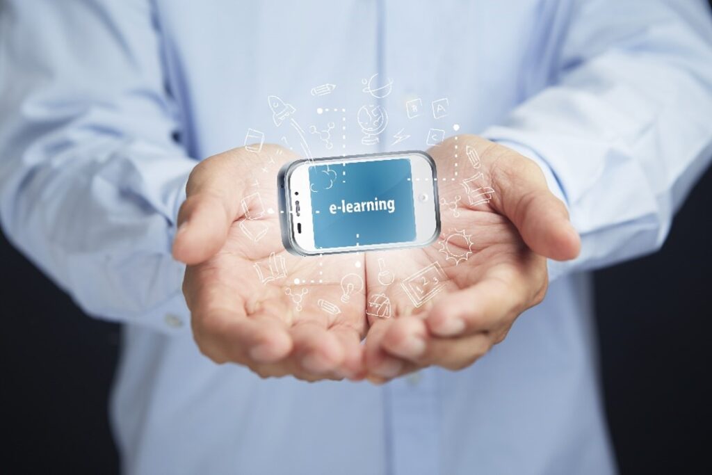 torso and arms of person wearing light blue button-down shirt with open hands facing up holding mobile phone with “e-learning” on the blue screen, with small white graphics coming out of the screen