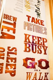 Tall and narrow poster with menu at the top and slogans in large fonts at the bottom. Background of poster is light beige, text is orange-brown. Slogans are a mix of serif and sans-serif bold fonts and include: Brew Tea, Take Pictures, Keep Busy, Go for a Walk. One starts with Sleep but the rest is cut off