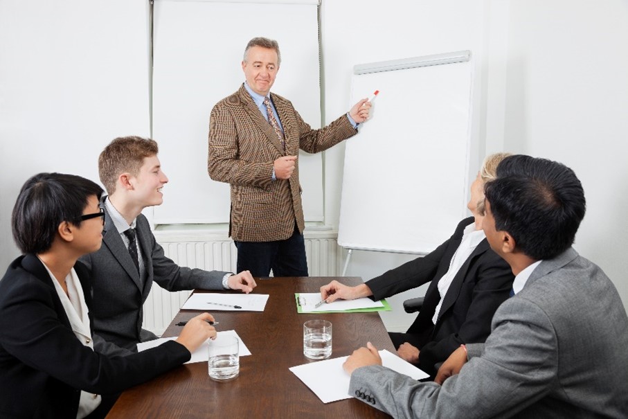 the same man as in earlier photo, but now he has a smile on his face. He is standing at a whiteboard on a white wall, presenting to a group of four people in suits (two women, two men) sitting at a dark brown wooden table with papers and glasses of water