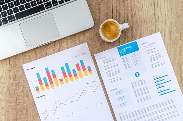 A silver laptop, two documents, and white mug of coffee are on a medium-brown woodgrain desk. One document shows Samantha Black’s resume, which is divided neatly into blue and black text sections with surrounding empty space. The second document is to the left, showing a multi-colored bar graph (blue, red, and yellow-orange) and a line graph with two lines colored green-grey and dark red.