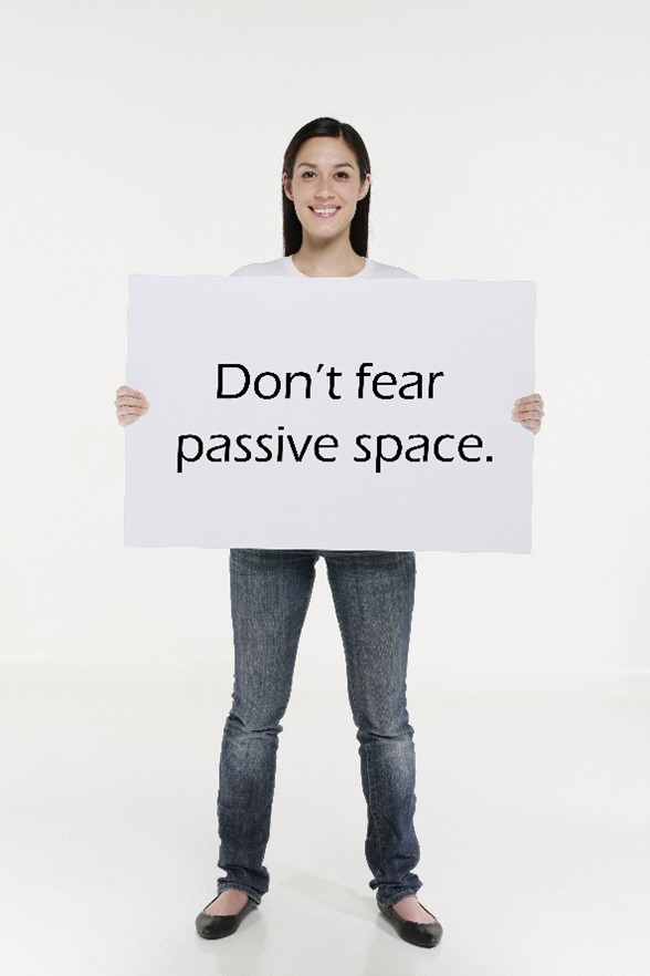 A smiling woman with dark, long, straight hair, wearing white t-shirt, medium-wash jeans, and black flats is holding a white sign that reads, “Don’t fear passive space.” The text is centered with lots of space around it, and she is standing in front of a white background and on a white floor